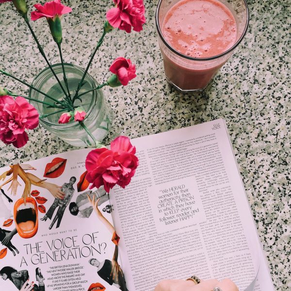 A top-down view of a magazine on a wooden table, surrounded by delicate flowers in soft tones and a vibrant pink smoothie in a glass, creating a fresh and inviting scene for implementing a DIY PR strategy.